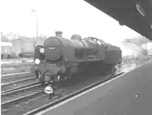 Side-and-front view of the rebuilt form of the locomotive, running without a train through a railway station. The locomotive is of conventional appearance, with a tender and has no side watertanks obscuring the boiler. Smoke deflectors are fitted at the front of the locomotive.