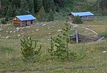 Two log cabins with metal roofs in the distance down a rolling slope. The remains of another are partially visible closer to the camera