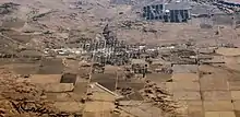 This is a photo taken from an airplane. The photo shows a city flanked by greenery, desert, and terrain patches. In the middle, the city is gridded, crisscrossed by streets. A pair of perpendicular roads, possibly highways, cross at the city's heart.