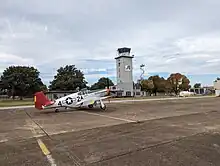 A grey, multi-story airport control tower. A P-51 mustang plane is parked in the foreground.