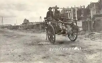 Two men in sombreros riding in a donkey-cart with a line of feet sticking out the back.  They are riding down a dirt street away from the camera, with a line of buildings on the right.  Dated 5/15/1911.