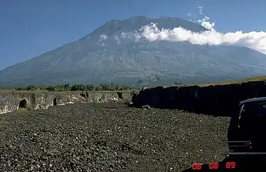 A photograph depicting a blue sky at the top, a grey mountain range in the middle, white clouds in front of the mountain range, and a rocky terrain at the bottom.