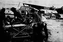 A man in a stretcher being taken to an ambulance, with damaged buildings in the background behind him.