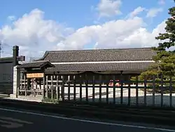 A wide wooden building with a large roof beyond a wooden fence.