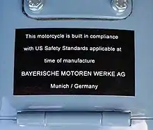 Black placard mounted on a light blue fuel tank, which reads "This motorcycle is built in compliance with US safety standards applicable at the time of manufacture BAYERISCHEN MOTOREN WERKE AG Munich / Germany"