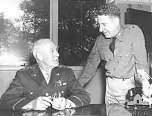 A man in uniform sits at a desk. He wears three stars and wings on his breast. Another, in a similar uniform but wearing only one star, leans over.