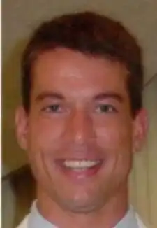 A smiling clean-shaven young man with short dark hair wearing a white shirt and silvery tie against a blue background