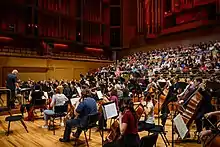 An orchestra sits on stage in a large hall. Behind them are choristers in the stands. There is a large pipe organ at the rear.