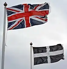 The Union and Cornish flags fluttering in the wind, against a grey, cloudy sky.