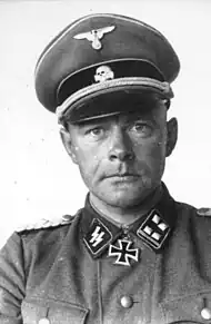 A man wearing a military uniform, peaked cap and a neck order in the shape of a cross. His cap has an emblem in shape of a human skull and crossed bones.