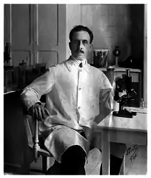 Black and white photo of Charlos Chagas, in his lab coat, sitting next to his microscope and surrounded by flasks and jars