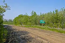 A muddy road with deep tire ruts and puddles on the left, amid a wooded area. At the left is a white-on-green sign with "Winter Ice ... Maximum Weight ... 000 kg" written on it. In the distance is some water reflecting evergreen trees across it.