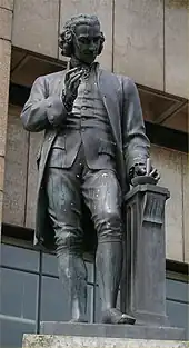 Statue of a man with a mortar and pestle in his left hand and his right hand upraised, holding a lens