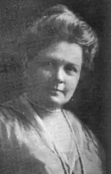 A middle-aged white woman, wearing her hair in a bouffant updo, and a strand of pearls over a satiny dress