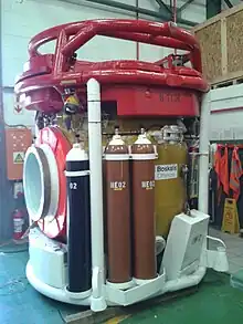 Exterior view of a closed bell, showing the side door to the left, with a 50-litre oxygen cylinder and two 50-litre heliox cylinders mounted to the frame to the side of the door.