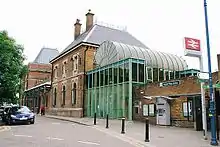 Photograph showing the older brickwork main station building with a green metal extension (now demolished) on the side that was formerly used as the ticket office.