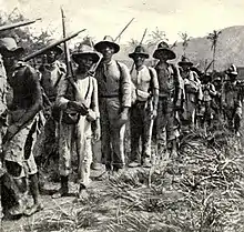 A column of Cuban Liberation Army soldiers marching. Most of the combatants are Afro-Cuban, wear torn clothing, and carry old rifles.