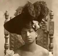 sepiatone headshot of a young woman sitting in a wooden chair, she is wearing a large black hat with elaborate feathers in it