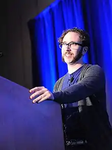 A man wearing a blue shirt and grey sweater looking to the left. He is standing behind a podium.