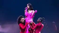 Dua Lipa performing in a pink catsuit being lifted by dancers in red outfits