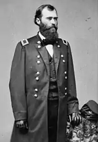 A white man with a full, bushy beard standing with his left hand resting on a table beside him; a slouch cap is on the table. He is wearing dark gloves, a long unbuttoned military jacket over a vest, and a bow tie.