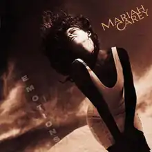 A brunette woman in a white low-cut minidress standing in front of a dark, sepia-toned desert-like background, hair flailing upward in the wind, the words "MARIAH CAREY" on the upper right hand side of the artwork and the word "EMOTIONS" vertically spelled out from the top down towards the bottom left hand side of the artwork.