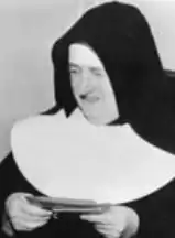 An older white woman in a black and white nun's habit, holding a small book in her hands.