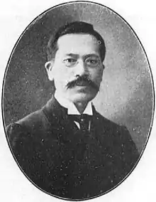 Black and white portrait photograph of Tasuku Harada in 1913. He is wearing a suit and glasses. He is looking directly into the camera. He has a large moustache.