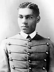 Cadet Henry O. Flipper in his West Point cadet uniform. It has three larger round brass buttons left, middle and right showing five rows. The buttons are interconnected left to right and vice versa by decorative thread. He is wearing a starched white collar and no tie. He is a lighter-colored African American with plated corn rows of neatly done hair. He is facing the camera and looking to the left of the viewer.