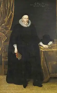 A tall elderly man with a beard, wearing long black robes and a large white ruff.  He is standing with a fan in his right hand and with his left hand resting on books on a table.