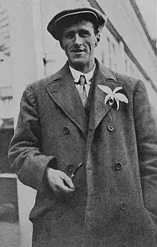 Picture of Herbert Whitley facing towards the camera, holding a tobacco pipe in his right hand down near his waist, and with an exotic flower in his left lapel