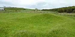 The ruins of the Hilton of Cadboll Chapel, which look like a series of gentle banks. In the background, directly in front of the viewer, stands the Hilton of Cadboll stone. It is grey and rectangular.