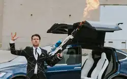 Jason Parsons wearing a white shirt and black suit with crisscross stripes, standing in front of a Model X Tesla, holding a Boring company flamethrower in one hand, with the other hand up in a rock symbol.