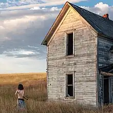 Jane Remover stands alone in a field with her back towards the camera as she faces the remnants of a dilapidated house.