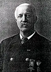 Head and shoulders of a white man with mustache wearing a double-breasted suit coat over a shirt and tie. Two medals are pinned to the coat's left breast.