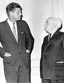 A candid portrait of Kennedy and Ben-Gurion standing facing each other, dressed formally, Kennedy with his hands in his suit jacket pockets.