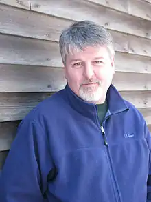 A portrait photograph of a white man in a blue jacket; he has greying hair on both his head and face and is looking into the camera with a neutral expression.