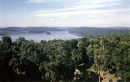 View across forest towards a lake surrounded by low, forested hills. Two small islands are set close to the far shore. There is a small clearing in the immediate foreground. On the left side of the far shore is a wide grassy region set against the forested hills.