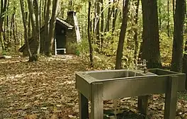 A metal sink with running water supported by wooden beams in the foreground, a wooden open shelter with a stone chimney amidst the trees in the background