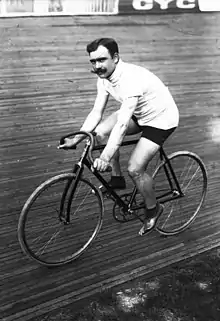 A black-and-white photograph of a man with three-colored sweater and shorts with a moustache sitting on a bicycle.