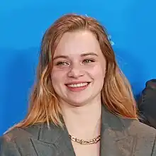 Luna Wedler wearing a grey jacket with lapels and a gold necklace, grinning and looking right of camera