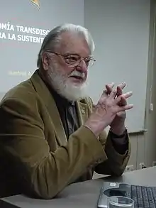 Manfred Max-Neef sits at a table near a notebook computer. On the wall behind him is a slide from a presentation.