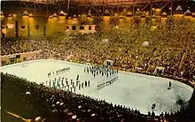 Far view of an ice hockey rink, with ice hockey players and a band standing at its centre. Spectators seated around the rink watch.