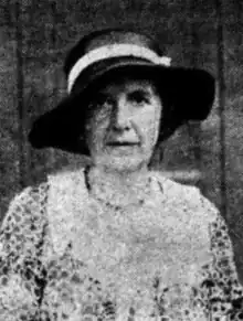 A middle-aged white woman, wearing a dark floppy hat and a print dress with a lace yoke detail