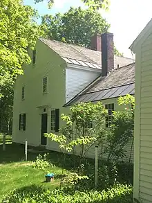 Image of the front of the white house, showing green front door and green window shutters. The lawn is bright green grass in front of the structure.