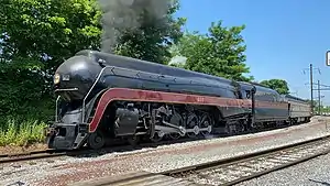 A black streamlined steam locomotive pulling an observation car with the trees and overhead lines in the background