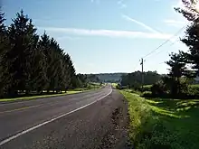 A two-lane highway heads through an area containing open fields and areas of trees. A large, tree-covered ridge is in the distant background.