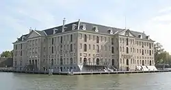 Light grey building with a dark grey roof surrounded by water