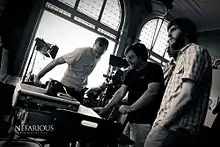 A black-and-white photograph of four men standing in a room filled with equipment including a videocamera, a speaker, and a spotlight