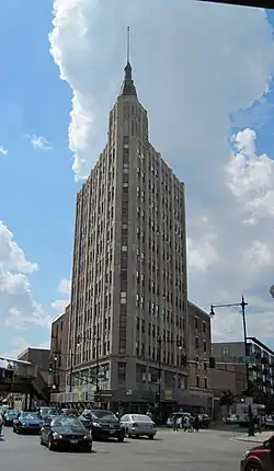 An art-deco skyscraper on an angled lot, with a pentagonal cupola and spire, viewed from the angled end. Significant portions of the street and automobile traffic are visible, as is the auxiliary exit of Damen Avenue on the viewer's extreme left overlying North Avenue.